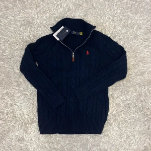 Mörkblå stickad tröja från Ralph Lauren - Säljer en snygg mörkblå stickad tröja från Ralph Lauren med dragkedja och röd broderad logga på bröstet. Tröjan har en ribbad krage och långa ärmar, perfekt för kyligare dagar. Passar både till vardags och mer uppklädda tillfällen.