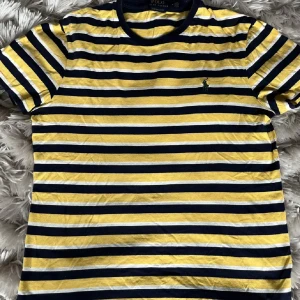 Randig t-shirt från Polo Ralph Lauren - Snygg randig t-shirt från Polo Ralph Lauren i gult och blått. Den har en broderad logga på bröstet och är kortärmad med en rund halsringning. Perfekt för en avslappnad stil.