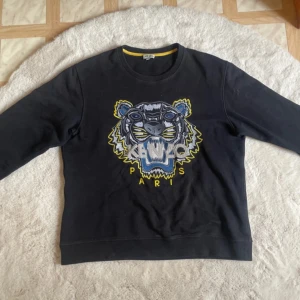 Svart sweatshirt från Kenzo med tigerbroderi - Säljer en svart sweatshirt från Kenzo med ett stort, färgglatt tigerbroderi på framsidan. Tröjan har rund hals och långa ärmar. Perfekt för en stilren och trendig look. 🐯