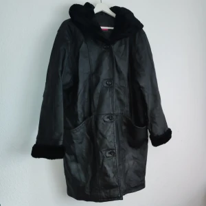 Svart skinnjacka med huva, for women - Snygg svart skinnjacka med huva och knappar framtill. Jackan har långa ärmar och är fodrad för extra värme. Perfekt för kyligare dagar med sin stilrena design och praktiska fickor.