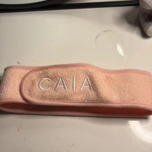 Rosa hårband från CAIA - Säljer ett mjukt rosa diadem från CAIA med broderad logga framtill. Perfekt för att hålla håret borta vid sminkning eller ansiktsvård. Justerbar passform med kardborreband. Aldrig använd.köpt för 95kr