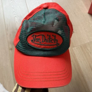 Röd och grön keps från Von Dutch - Snygg röd keps från Von Dutch med grönt nät och broderad logga framtill. Perfekt för en avslappnad stil. Justerbar passform för optimal komfort.