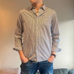 Tommy hilfiger skjorta - Snygg randig skjorta i blått och vitt med långa ärmar och klassisk krage. Skjortan har knappar framtill och en liten broderad detalj på bröstet. Perfekt för en stilren look. Tommy hilfiger 