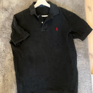 Svart pikétröja från Ralph Lauren - Säljer en klassisk svart pikétröja från Ralph Lauren med den ikoniska röda broderade loggan på bröstet. Tröjan är tillverkad i 100% bomull och har korta ärmar samt knappar vid kragen. Perfekt för en stilren look.