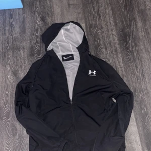 Svart vindjacka från Under Armour - Säljer en svart vindjacka från Under Armour med dragkedja och huva. Jackan har en lös passform och är perfekt för blåsiga dagar. Den är tillverkad i ett lätt syntetmaterial och har ett diskret märke på bröstet.