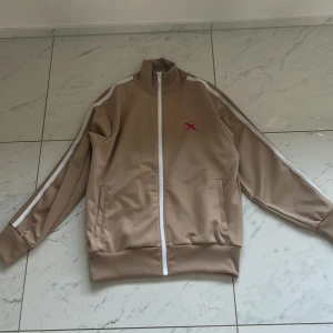 Beige track jacket från Axel Arigato - Snygg beige track jacket från Axel Arigato med vita detaljer och en röd broderad logga på bröstet. Jackan har en dragkedja framtill och vita ränder längs ärmarna. Perfekt för en stilren och sportig look.