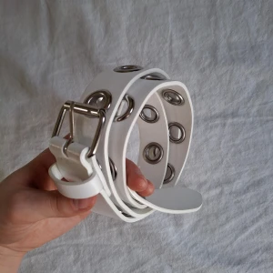 Vitt bälte med metallringar - Snyggt vitt bälte med stora metallringar längs hela längden. Perfekt för att ge en edgy touch till din outfit. Bältet har ett klassiskt spänne i silverfärgad metall och passar alla storlekar.