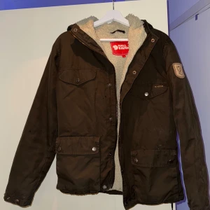 Fjällräven jacka - Säljer en brun jacka från Fjällräven med mysigt fleecefoder. Jackan har dragkedja och knappar framtill samt flera praktiska fickor. Perfekt för kyliga dagar.