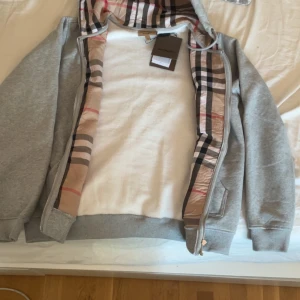 Grå huvtröja från Burberry - Säljer en stilren grå huvtröja från Burberry med klassiskt rutigt mönster på insidan av huvan. Tröjan har dragkedja och är perfekt för en avslappnad look. Märkets logga finns på etiketten och den är i storlek L.