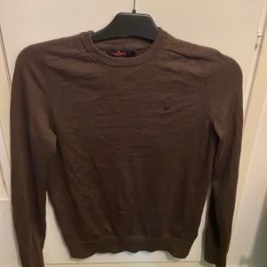 Brun tröja i merinoull från Morris - Säljer en brun tröja från Morris i 100% merinoull. Tröjan har en rund halsringning och ett diskret broderat emblem på bröstet. Perfekt för kyligare dagar.