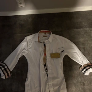 Vit skjorta från Burberry - Snygg vit skjorta från Burberry med klassiska rutor på insidan av kragen och ärmsluten. Skjortan har långa ärmar och en broderad logga på bröstet. Perfekt för en fet kväll med boysen🤩🤩💫⭐️(priset är inte hugget i sten) fick den i present av min farfar så vet inte nu priset😅