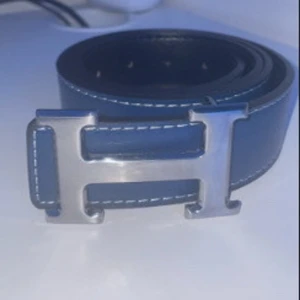 Blått bälte med silverfärgat spänne - Snyggt blått bälte med ett stort silverfärgat H-spänne. Tillverkat i skinn med synliga sömmar längs kanterna. Perfekt för att ge en stilren touch till din outfit.