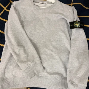 Grå sweatshirt från Stone Island - Säljer en stilren grå sweatshirt från Stone Island med deras ikoniska märke på ärmen. Tröjan har en rund halsringning och långa ärmar, perfekt för en avslappnad look. Passar bra till både jeans och joggers. Hör av dig vid intresse!