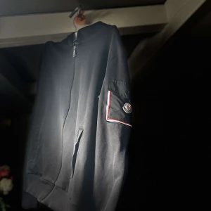 Svart monclertröja med dragkedja  - Snygg svart tröja från Moncler med dragkedja framtill. Tröjan har en ribbad krage och muddar, samt en broderad logga på ärmen. Perfekt för en stilren look.