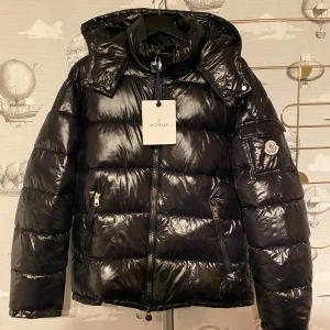 Svart pufferjacka från Moncler - Snygg svart pufferjacka från Moncler med glansig finish. Jackan har en dragkedja framtill och en avtagbar huva. Den är långärmad och har Monclers logotyp på ärmen. Perfekt för kalla dagar med sin quiltade design.
