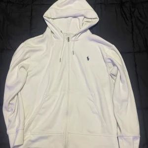 Vit hoodie från Ralph Lauren - Säljer en stilren vit hoodie från Ralph Lauren med dragkedja och klassisk logga på bröstet. Tröjan har långa ärmar och en praktisk huva med snörning. Perfekt för en avslappnad look.