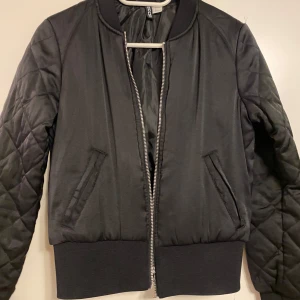 Svart bomberjacka från Divided - Snygg svart bomberjacka från Divided med quiltade ärmar och dragkedja framtill. Jackan har ribbade muddar vid ärmslut och nederkant. 