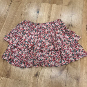 Blommig volangkjol från Zara - Snygg kortkjol med blommigt mönster i rosa, rött och grönt. Kjolen har volanger och en dragkedja i sidan. Den har också insydda shorts under som gör den mer rörlig.