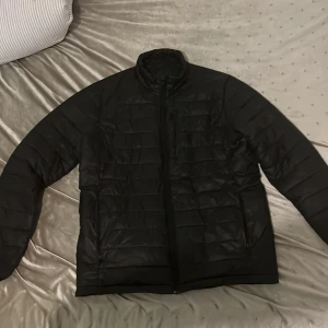 Svart pufferjacka med Thermolite - Säljer en svart pufferjacka med Thermolite-isolering. Jackan har en dragkedja framtill och är quiltad för extra värme. Perfekt för kyliga dagar. Den är långärmad och har en stilren design.