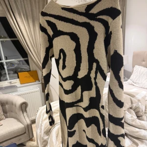 Beige och svart mönstrad klänning - Snygg långärmad klänning i beige och svart med ett unikt abstrakt mönster. Perfekt för att sticka ut med stil. Klänningen har en bekväm passform och är tillverkad i ett mjukt material.