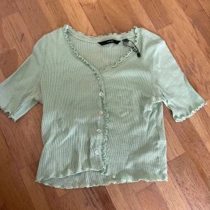 Ljusgrön ribbad topp från Vero Moda - Säljer en ljusgrön ribbad topp från Vero Moda med volangkant längs v-ringningen och knäppning framtill. Toppen har korta ärmar och en croppad passform. Den är i bra skick och perfekt till sommaren.🍀🌿