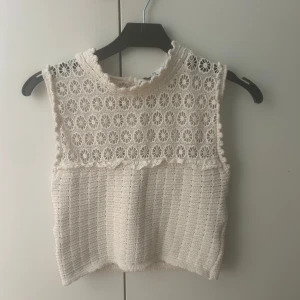 Vit spetsdetaljerad topp från Zara - Säljer en vit croppad topp från Zara med vackra spetsdetaljer upptill och virkad struktur nedtill. Toppen är ärmlös och har en hög hals med fina blommönster i spetsen. Perfekt för sommardagar och att styla med jeans eller kjol.