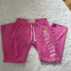 Rosa lågmidjade mjukisbyxor från Hollister - Säljer ett par rosa lågmidjade mjukisbyxor från Hollister med vit dragsko i midjan och tryckt text längs benet. Byxorna har normal passform och är perfekta för en avslappnad stil. Mjukt bomullsmaterial och resår i midjan. Jag är 166 cm lång! Inga defekter💗💗