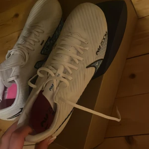 Vita Nike Air Zoom fotbollsskor - Säljer ett par vita Nike Air Zoom fotbollsskor med blå detaljer och rosa innersula. Skorna har snörning och en sportig design med svart Nike-logga på sidan. Perfekta för dig som vill ha lätta och snygga skor på planen. Jag har storlek 38-39 men man ska alltid ha lite större skor när det gäller fotbollsskor eftersom dom brukar vara väldigt små i storlek 