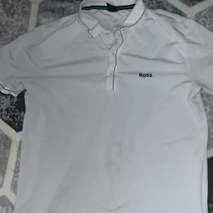 Vit slim fit t-shirt från BOSS - Säljer en vit t-shirt från BOSS med klassisk krage och knappar framtill. T-shirten har en diskret BOSS-logga på bröstet och är i slim fit-modell. Perfekt för en stilren och enkel look.