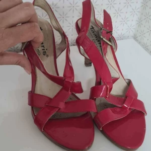 Röda sandaler med klack från Tamaris - Säljer ett par röda sandaler från Tamaris med öppen tå och snygga korsade remmar. Skorna har justerbart spänne vid ankeln och en medelhög klack. Perfekta för dig som vill ha en färgklick till outfiten!