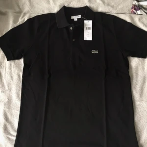 Svart pikétröja från Lacoste - Klassisk svart pikétröja från Lacoste med den ikoniska gröna krokodilloggan på bröstet. Tröjan har korta ärmar, krage och knäppning med två knappar framtill. Perfekt för en stilren och sportig look.