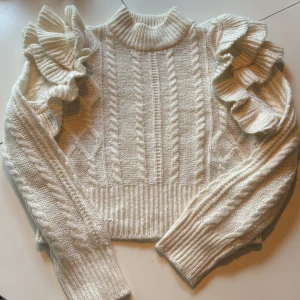 Vit stickad tröja  från Gina Tricot - Supersöt vit stickad tröja från Gina Tricot med volangdetaljer på axlarna och kabelstickat mönster. Tröjan har  långa ärmar med ribbade muddar. Perfekt för att lyfta din outfit!