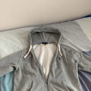 Grå hoodie från Polo Ralph Lauren - Säljer en grå hoodie från Polo Ralph Lauren med dragkedja och vit fleecefoder på insidan. Tröjan har huva med vita snören och två fickor framtill. Klassisk liten logga broderad på bröstet.