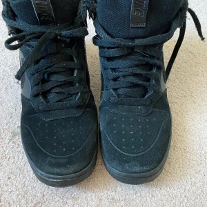 Svarta Nike sneakers high-top - Säljer ett par svarta Nike high-top sneakers med snörning och klassisk swoosh på sidan. Skorna har en platt sula och är tillverkade i syntetmaterial och skinn. Perfekta för dig som gillar streetstyle och vill ha en stilren look. Fodrade inuti perfekt till vintern 