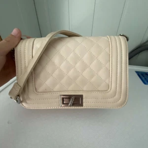 Beige quiltad axelväska med kedja - Säljer en beige axelväska med quiltat mönster och silverfärgad kedja. Väskan har ett stilrent spänne framtill och är tillverkad i syntetmaterial. Perfekt för dig som vill ha en snygg och trendig accessoar till din outfit. Fås billigare vid snabb affär 