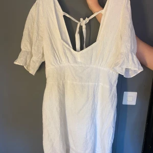 Studentklänning vit klänning - Säljer en fin vit klänning från Nelly med broderade blommor, v-ringning och knytning i ryggen. Klänningen har korta puffärmar och är tillverkad i ett lätt bomullstyg. Perfekt för studenten och sommarens alla tillfällen!