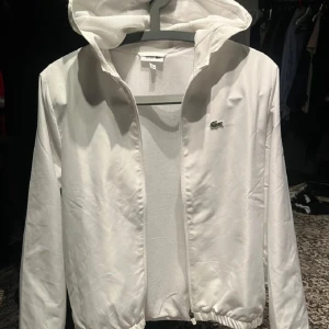 Vit vindjacka från Lacoste - Snygg vit vindjacka från Lacoste med huva och dragkedja. Den har bara använts engång.         Jackan har det klassiska krokodilloggan på. Stilig och prefekt för vår, sommar och höst. 