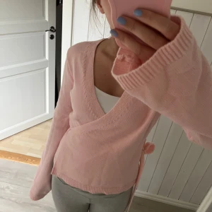 Ljusrosa omlottkofta med rosett - Säljer en söt ljusrosa omlottkofta med knytband och rosett i sidan. Tröjan har lång ärm och är stickad i ett mjukt material. Helt i nyskick då den aldrig kommit till användning.