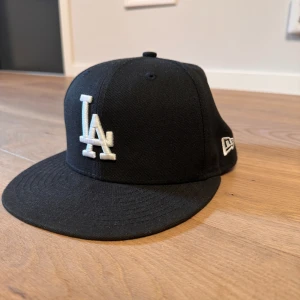 Svart LA-keps från New Era 59FIFTY - Snygg svart keps från New Era 59FIFTY med LA-logga broderad framtill i vitt. Klassisk modell med platt skärm och MLB-logga baktill. Storlek 7 1/8 (56,8 cm). Perfekt för dig som gillar sportig stil och streetwear.