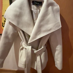 Beige kappa med bälte från Vero Moda - Snygg beige kappa från Vero Moda med stor krage och knytbälte i midjan. Jackan har lång ärm och en klassisk, elegant look som passar perfekt till sommar (med tanke på Sveriges sommar väder där de kan bli regnit o kallt) och vår. Enkel att matcha med olika outfits. Jackan är inte andvänd. 