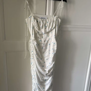 Vit blommig klänning från Adoore - Supersöt vit klänning från Adoore med små blå blommor, smala axelband och rynkade detaljer längs sidan. Klänningen har en feminin siluett och är perfekt för sommarens alla tillfällen. Axelbandsklänning med dragsko vid bysten.