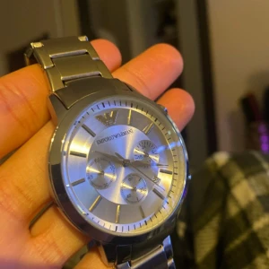 Silverfärgad klocka från Emporio Armani - Stilren herrklocka i silver med metallarmband och rund urtavla. Klockan har tre mindre urtavlor och tydliga indexmarkeringar. Emporio Armani-loggan syns högst upp på urtavlan.