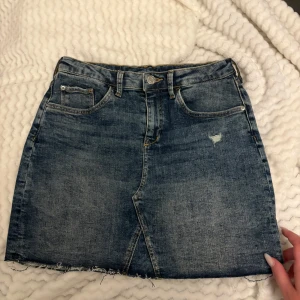 Mörkblå jeanskjol med rå kant - Säljer en mörkblå jeanskjol med klassisk femficksdesign och rå, fransig nederkant. Kjolen har bälteshällor, knapp och dragkedja framtill samt en liten sliten detalj på framsidan. Perfekt till vardags med en t-shirt eller hoodie. Passar XXS och XS! Även justerbar i midjan.