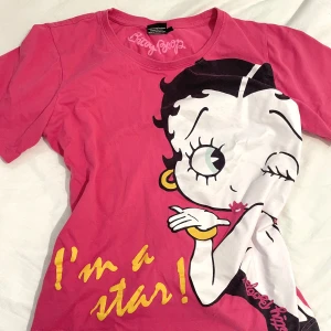 Rosa Betty Boop t-shirt med tryck - Säljer en rosa t-shirt från Betty Boop med stort tryck av Betty Boop och texten 'I'm a star!' i gult framtill. T-shirten är i 100% bomull och har en klassisk passform med korta ärmar. 