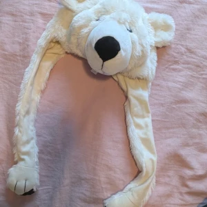 Vit isbjörnsmössa med långa öronlappar - Rolig och mjuk mössa i form av en isbjörn med långa öronlappar. Mössan är vit och har ett stort svart nosparti. Perfekt för att sticka ut eller hålla sig varm under vintern.