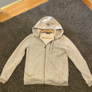 Ljusgrå hoodie från Sail Racing - Säljer en ljusgrå hoodie från Sail Racing med dragkedja framtill och huva. Tröjan har två fickor på sidorna och diskret logga på bröstet. Perfekt för en avslappnad stil och enkel att matcha med det mesta.