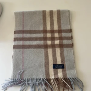 Rutig halsduk från Burberry i kashmir - Säljer en klassisk rutig halsduk från Burberry i 100% kashmir. Halsduken har beige, bruna och grå toner med fransar i ändarna och den ikoniska Burberry-loggan. Perfekt accessoar för att lyfta din outfit. Scick 9/10 då knappt använd priset är ej hugget i sten och tveka inte på att höra av er vid frågor