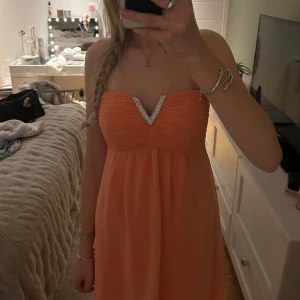 Axelbandslös korallfärgad klänning med strassdetalj - Säljer en axelbandslös korallfärgad klänning med v-ringning dekorerad med strass. Klänningen har plisserad överdel och faller lätt i chiffongliknande material. Perfekt för festliga tillfällen. Hålen gjorde jag med flit pågrund av att jag skulle sy den kortare men har inte orkat, färgen är lite aprikos 