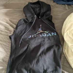 Svart Jordan hoodie med regnbågslogga - Säljer en svart hoodie från Jordan med en stor regnbågsfärgad logga och text på bröstet. Tröjan har huva med dragsko och en stor magficka. Perfekt för dig som gillar sportig och avslappnad stil.