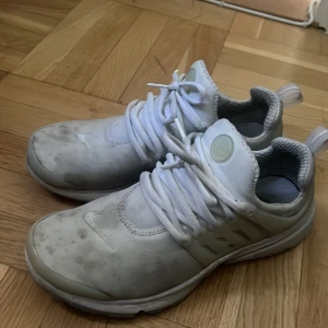 Vita Nike Presto sneakers - Säljer ett par vita Nike Presto sneakers med rund tå och platt sula. Skorna har snörning och är tillverkade i syntetmaterial med mesh-detaljer för extra andningsförmåga. Perfekta för dig som gillar en sportig och clean stil.